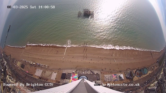 Aerial view of a beach and pier in Brighton, England, taken on a sunny day. The image shows the sandy shoreline with people walking along the beach, and gentle waves lapping at the shore. The ocean is a shimmering blue-green color, reflecting sunlight. In the distance, part of the pier is visible extending into the water. Below the beach, colorful beach umbrellas and structures can be seen, alongside buildings along the promenade.