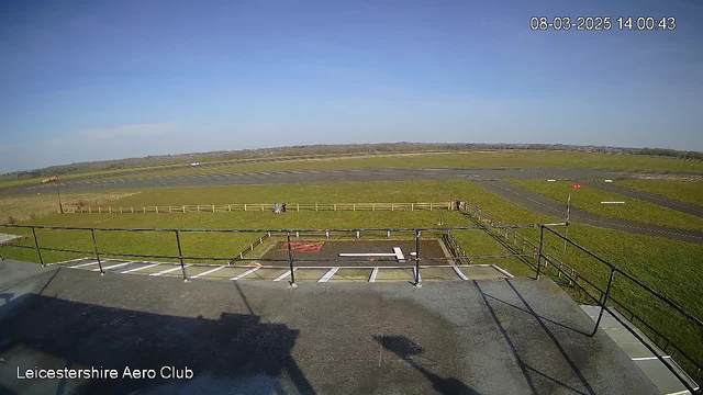 A clear blue sky stretches above a grassy airfield. In the foreground, a balcony with railing overlooks the airfield. There are two people walking on the grass, and a small area marked for a helicopter landing pad is visible below. In the distance, a single-engine aircraft is positioned on the runway, which is mostly paved with some grassy areas surrounding it. There are a few wind indicators on the field. The scene is bright and sunny, conveying a calm, open atmosphere.