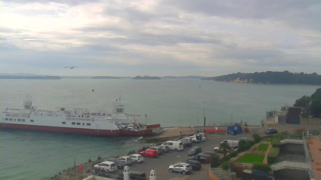 A coastal scene featuring a large ferry docked near a shoreline. The ferry is white with a red bottom and has vehicles on board. Nearby, there is a parking area filled with cars of various colors. The water is calm, reflecting the overcast sky filled with gray clouds and a few distant islands visible in the background. A seagull is flying above the ferry, and vegetation and structures can be seen on the land to the right.