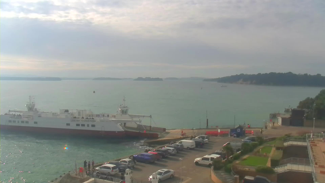 A large ferry is docked at a harbor, with people visible on board. The body of water is calm, reflecting the sky, which is partly cloudy. In the foreground, there is a parking lot filled with various cars, and a small coastline with greenery and buildings can be seen on the right side. The horizon features distant trees and landmasses.