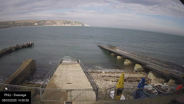 A coastal scene shows a pier extending into the water from a concrete ramp. The ramp leads from a rocky shoreline with pebbles and patches of seaweed. On the right, two kayaks are parked near a few boats. The ocean is visible in the background, with gentle waves and a cloudy sky overhead. The white cliffs are faintly seen in the distance along the shoreline.