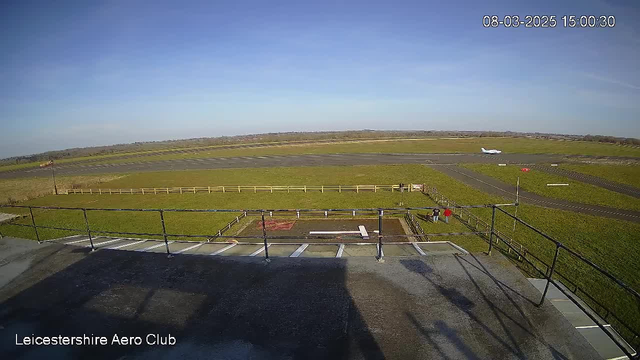 The image shows an outdoor view from a high vantage point at Leicester Aero Club. In the foreground, a metal railing outlines a rooftop area with a solid surface. Below, a grassy field extends toward a runway where a small airplane is taking off. To the right, a group of people is gathered near wooden fencing. The sky is clear and blue, indicating a bright day. The time and date are displayed in the top corner of the image.