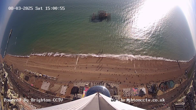 An aerial view of a beach with golden sand stretched along the shoreline, where small waves gently wash up. In the distance, there's a wooden pier extending into the water. Several people are walking along the beach, and some are enjoying activities nearby. A colorful amusement park is visible on the left side of the image, with various attractions and a swimming pool. Above the beach, the ocean appears calm and reflects sunlight, creating a shimmering effect. The scene is bright and clear, depicting a sunny day.