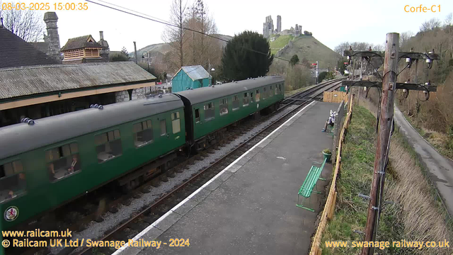 A green train carriage is passing through a railway station. In the background, there are green hills and a castle ruins on a slope. The station platform has a green bench and a wooden fence. A telephone pole with wires is visible on the right side of the image. Trees and a building with a sloped roof are in the upper part of the scene. The sky is bright and clear.