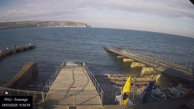 A coastal scene featuring a rocky shoreline with two wooden piers extending into the sea. The left pier has several individuals standing on it, while the right pier leads towards a concrete platform. In the foreground, there are small boats, one yellow and one blue, positioned on the shore amidst scattered rocks. The water appears calm, and the distant cliffs are visible, under a mostly clear sky. The image is timestamped, indicating the date and time.