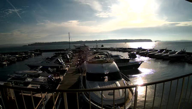 A photo of a marina during the day, featuring numerous boats docked in the water. The surface of the water reflects sunlight, creating a shimmering effect. In the background, there are hills and a clear sky with scattered clouds. The image captures the tranquility of a waterfront setting.