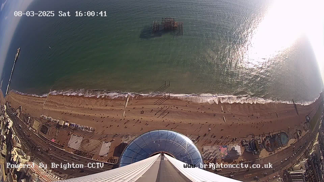 Aerial view of a beach with golden sand and gentle waves lapping at the shore. People walk along the beach, while a pier extends into the sea in the distance. An old, partially submerged structure is visible in the water. The scene is bright with sunlight reflecting on the surface of the ocean. In the foreground, a circular glass structure appears to be part of an observation deck. The sky is mostly clear, with a few clouds scattered across.