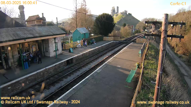 A view of Corfe Castle railway station showing a platform with several people waiting. To the left, there is a stone building with a sloped roof and large windows. A group of children is gathered near the entrance of the building. On the platform, there are green benches and a sign indicating "WAY OUT." In the background, the ruins of Corfe Castle sit atop a hill, surrounded by trees and rolling landscape. The scene is illuminated by late afternoon sunlight. The tracks run parallel to the platform, extending into the distance.