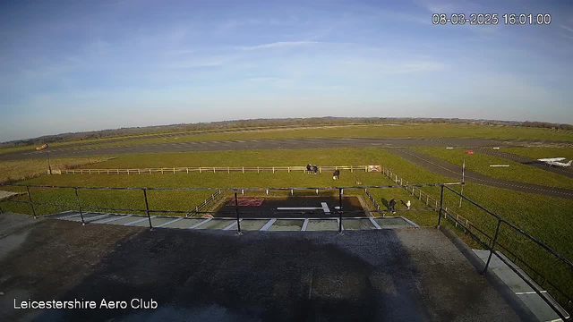 A wide view of an aerodrome on a clear day. In the foreground, a railing indicates the edge of a building. Below, there is a runway marked with a white strip, surrounded by green grass. A few people are scattered in the grass area, and a small red sign can be seen nearby. In the background, the runway extends into the distance, bordered by low hills under a blue sky with scattered clouds. The time and date indicate it is 16:01 on March 8, 2025.