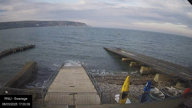 A coastal scene featuring a view of the sea with calm waves. In the foreground, there are stone steps leading down to the water's edge, flanked by a railing. To the left, a wooden pier extends into the water. There are several boats and yellow kayaks stored near the steps on a pebbly shoreline. In the distance, the coastline rises with cliffs and greenery, under a partly cloudy sky.