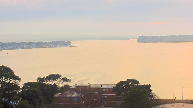 A tranquil view of a body of water at dawn, reflecting soft oranges and yellows from the rising sun. In the foreground, there are buildings with a red brick exterior and greenery from nearby trees. On the horizon, a shoreline is visible with boats dotting the water, while further back, gentle hills fade into a hazy sky.