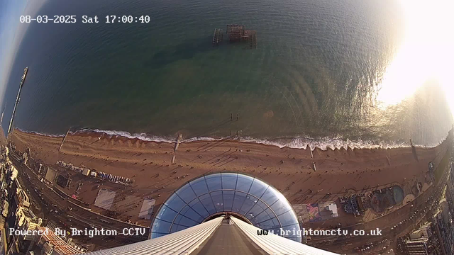 An aerial view from a high vantage point overlooking a sandy beach and the ocean. The beach is mostly empty, dotted with a few people near the shoreline. A structure resembling a pier is visible extending into the water, with another submerged structure in the ocean. The sea appears calm with gentle waves. In the foreground, a glass dome is visible, along with white railings and a pathway leading downward. The image also displays a timestamp in the upper left corner indicating the date and time.