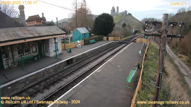 A view of a railway station. The foreground shows a platform with two green benches and a sign indicating "Way Out." On the left, there is a stone building with a sloped roof and several windows. To the right, a wooden fence encloses landscaped areas with a small tree and some greenery. In the background, there is a hill with what appears to be the ruins of a castle. The sky is cloudy, and the image captures an afternoon scene with soft lighting.