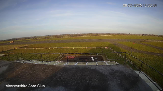 A wide view of an airfield under a clear blue sky. In the foreground, there is a flat rooftop with a railing on the left side. Below, a grassy field stretches out towards a paved runway that extends across the image. The runway is marked with white lines, and a red windsock is visible on the left, indicating wind direction. The background features distant trees and a horizon. The text at the bottom indicates the location as Leicestershire Aero Club, with a timestamp in the corner showing the date and time.