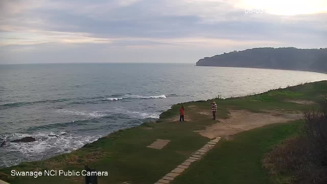 A scenic view of the coastline with calm waves gently lapping at the shore. In the foreground, two individuals are standing on a grassy area, one wearing a bright red top and the other in a checkered shirt. There are several stone pathways leading towards the water. In the background, a cliff rises along the coastline under a partly cloudy sky. The scene appears tranquil and natural, reflecting a peaceful outdoor setting.