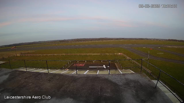 A wide view from a webcam at the Leicestershire Aero Club shows an expansive field with a small runway area. The ground is mostly green grass, with a few patches of dirt. A white wind sock is visible on the left, indicating wind direction. In the foreground, there is a fenced area with a red and white square marking on the surface. The sky above has soft shades of pink and blue, suggesting late afternoon or early evening light. The image conveys a sense of calmness typical of an airport setting.