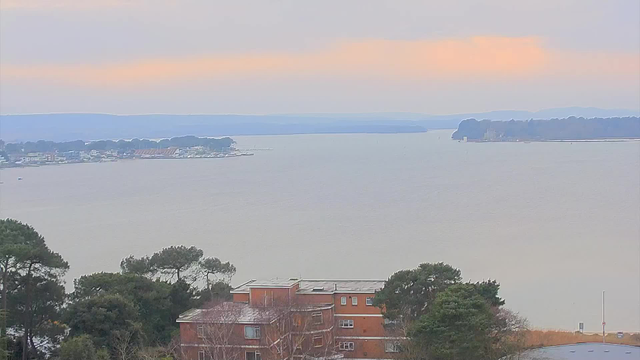 A view of a calm body of water under a cloudy sky, with soft hues of orange and pink near the horizon. In the foreground, there are trees with green foliage and a brick building with multiple windows. Across the water, a shoreline is visible with a mix of buildings and boats, leading into a distant mountainous landscape. The atmosphere is tranquil, suggesting a serene environment.