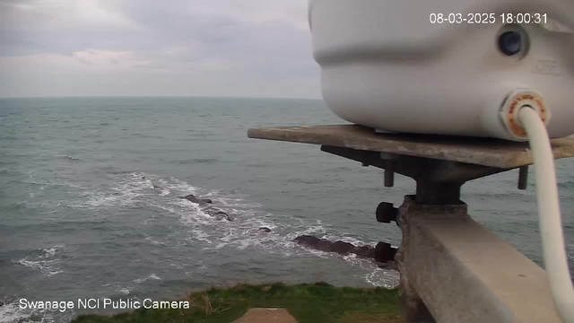 A view from a webcam overlooking the ocean. In the foreground, there is a white camera attached to a metal structure. Below the camera, grassy land meets the rugged shoreline. The ocean stretches out to the horizon, where the water appears calm with gentle waves. In the distance, there are visible rock formations just above the water's surface. The sky is overcast with clouds, suggesting a gray atmosphere.