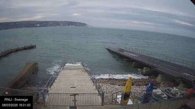 A view of a coastal area featuring a gray, cloudy sky. The ocean appears choppy with small waves lapping against a stone and concrete ramp leading down to the water. On the right, there is a wooden jetty extending into the sea. To the side, several kayaks in bright colors—yellow, red, and blue—are positioned on the shore near a railing. The landscape includes cliffs in the background, fading into the horizon. The scene is calm but has an overcast ambiance.