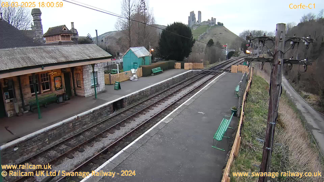 A train station platform is shown from a webcam perspective. The foreground features a stone wall and a paved area with several green benches. Beyond the benches, there are railway tracks running horizontally across the image. In the background, a small green building with a sloped roof stands beside a wooden fence. To the left, a sign reads "WAY OUT." In the distance, a castle ruin is visible situated on a hill, surrounded by trees and grassy landscape. The sky is gray, indicating overcast weather, and the time displayed in the corner reads 18:00:59 on March 8, 2025.