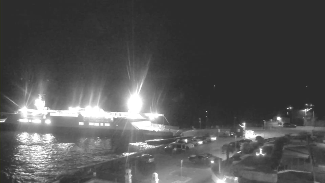 A brightly lit ferry is docked at a harbor during nighttime, surrounded by water that reflects the light. Several vehicles are parked along a nearby road, with some faintly visible lights in the background. The scene is predominantly dark, but the bright lights from the ferry and the surrounding area provide illumination.