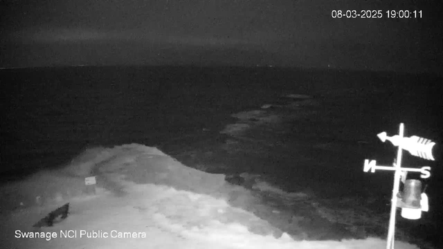 A black and white image of a seascape at dusk. The water appears dark and calm, with faint reflections. A rocky shoreline is visible in the foreground, leading into the ocean, where a light mist can be seen over the surface. In the lower right corner, there is a weather vane with directional indicators, and it is labeled "S" for south. The timestamp at the top shows the date and time as March 8, 2025, at 19:00:11.