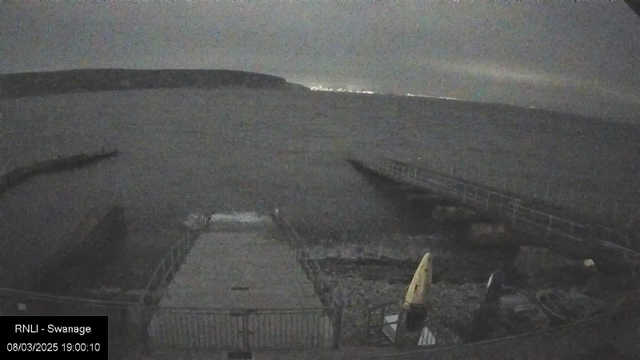 A dimly lit coastal scene with dark water extending into the distance. A partially visible pier juts out from the left side, and a small rocky area is present in the foreground. Two kayaks are positioned near the edge of the pier, one yellow and one darker in color. In the far background, faint lights can be seen along a distant shoreline, while overcast skies dominate the scene.