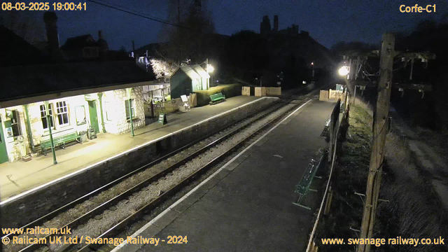 A dimly lit train station at night. There is a platform with a few green benches and a sign indicating an exit. The station building is visible on the left, with light illuminating part of its stone facade and windows. A wooden fence separates the platform from the area behind it. Several trees and structures are silhouetted against the dark sky. Two railway tracks run along the platform, leading into the distance. A streetlamp provides a warm glow in the scene.