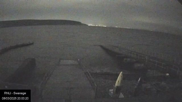 A dimly lit webcam image shows a serene body of water under an overcast sky. In the foreground, there is a wooden dock extending into the water, with a few small boats stored nearby. The distant shoreline is slightly illuminated, suggesting a town or village. The overall atmosphere is calm and quiet, reflecting a nighttime coastal scene.