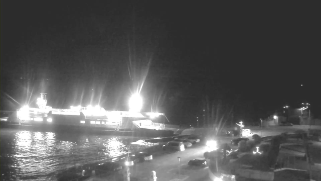 A brightly lit ferry docked at a dark waterfront. The water reflects some of the light from the ferry, and there are several cars parked along the shoreline. The scene is illuminated by lights from the ferry and nearby buildings, creating a contrast against the dark night sky.