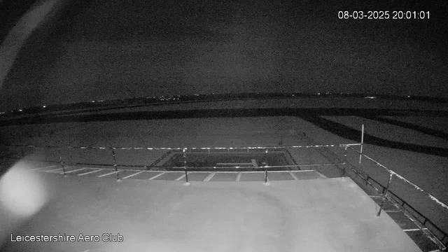 A nighttime webcam view of the Leicestershire Aero Club. The image is mostly dark, showing a paved area with parking spaces at the bottom, marked by white lines. In the background, there are faint lights visible along the horizon, indicating the presence of distant buildings or structures. The scene is framed by a railing along the edge. The overall ambiance is quiet and calm, typical of late evening settings.