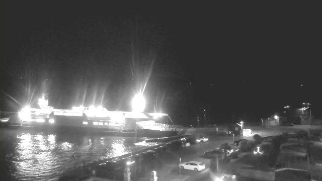 A brightly lit ferry docked at night, reflecting light on the water. Surrounding the ferry, there are parked vehicles along the pier, with faint outlines of buildings and lights on the shore in the background. The overall scene is dark, with the ferry being the primary source of illumination.