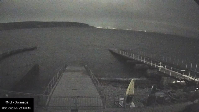 A dimly lit scene showing a calm body of water. In the foreground, there is a concrete jetty with metal railings. To the right, a wooden walkway extends into the water. On the left side, two kayaks are propped against a fence. The sky is overcast with little visible light, and a distant landmass is visible across the water.