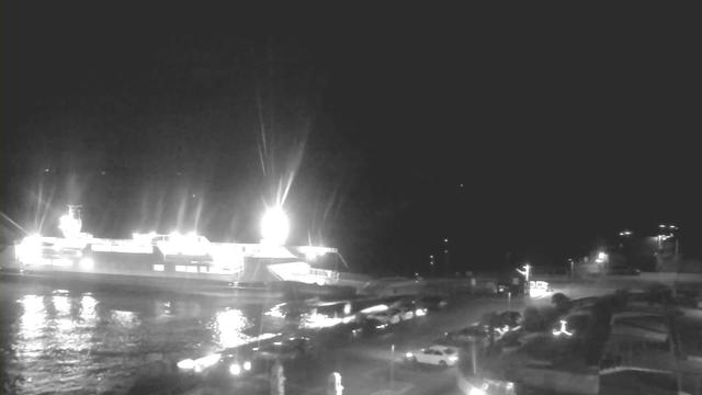 A dimly lit scene captures a large ferry docked at a wharf, illuminated by bright white lights. The water reflects these lights, creating a shimmering effect on the surface. In the foreground, there is a parking area with several vehicles, including some parked cars and a small bus. The background features a dark sky with faint distant lights, suggesting a coastal or harbor area at nighttime.