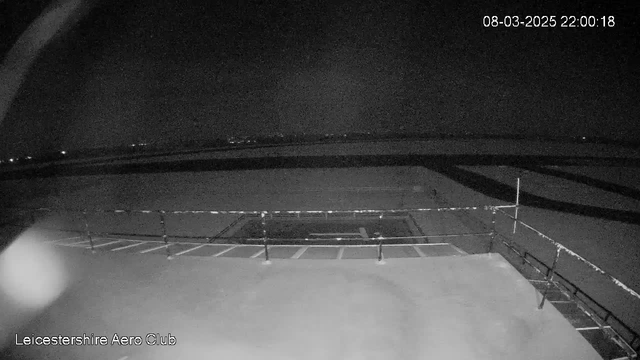 A dark scene captured by a webcam at the Leicestershire Aero Club. The image shows a flat, snowy surface likely representing a runway or tarmac area. There are faint outlines of markings on the surface, and a fence or railing is visible in the foreground. In the distance, some lights are barely visible, indicating a low level of ambient light, suggesting it's nighttime. The date and time display indicates it's March 8, 2025, at 10:00 PM.