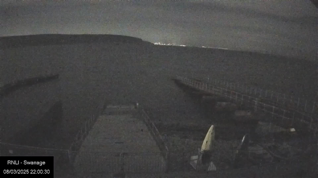 Nighttime view of a calm water surface with a faint horizon line in the distance. In the foreground, there is a wooden pier extending into the water. To the right, two small boats are partially visible on a concrete platform. The scene is primarily dark, with minimal light illuminating the distant shoreline.