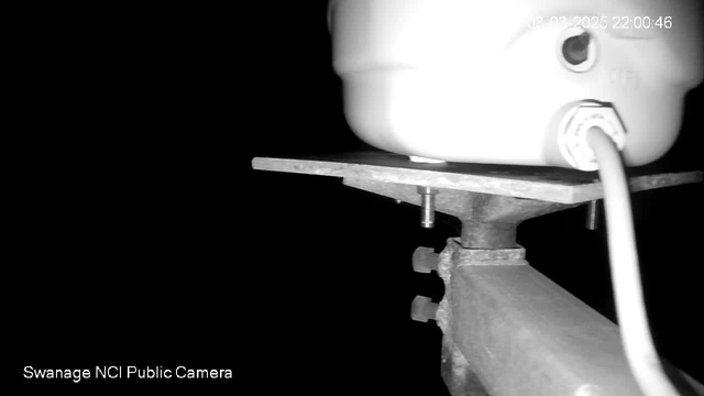 A close-up view of a white cylindrical object mounted on a flat surface, secured with screws. The background is dark with minimal visible detail. A power cable is connected to the object. The date and time display indicates the image was taken on March 8, 2025, at 22:00:46. A label at the bottom reads "Swanage NCI Public Camera."