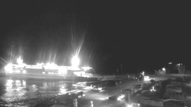 A night scene featuring a lit ferry docked at a pier, with bright lights illuminating the vessel. The water reflects the lights, creating a shimmering effect. Several vehicles are parked near the dock, and there are silhouettes of additional structures in the background, suggesting a coastal setting. The scene conveys a quiet, nighttime atmosphere with minimal movement visible.