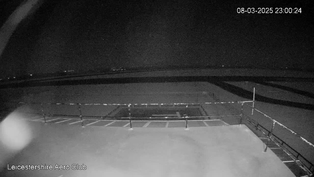 A black and white image of a nighttime scene at Leicestershire Aero Club. A flat surface is visible in the foreground, with a railing along one side. The background shows a dark sky with faint distant lights. The scene appears quiet and devoid of activity. A time stamp in the upper right corner indicates the date and time as March 8, 2025, at 23:00:24.