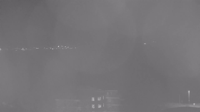 A monochrome image taken from a webcam shows a dimly lit scene at night. In the foreground, a few buildings can be seen, with some windows illuminated. The background features a faintly lit horizon where distant lights appear, possibly representing streetlights or buildings. The overall atmosphere is dark and hazy, with limited visibility due to a low light condition.