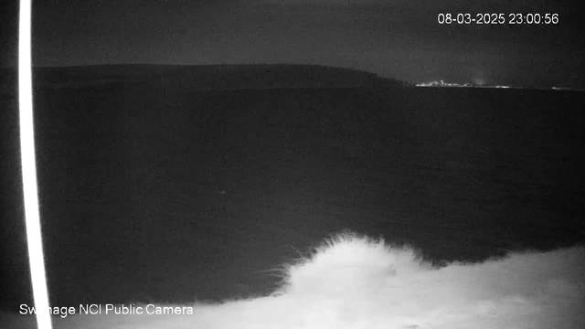 A dimly lit view of the ocean at night, with a dark horizon where the water meets the sky. The foreground shows white frothy waves breaking against the shore. There is a faint glow of lights in the distance, hinting at a coastal town or city. A vertical white line appears on the left side, possibly from the webcam lens, with a timestamp displayed in the upper right corner.