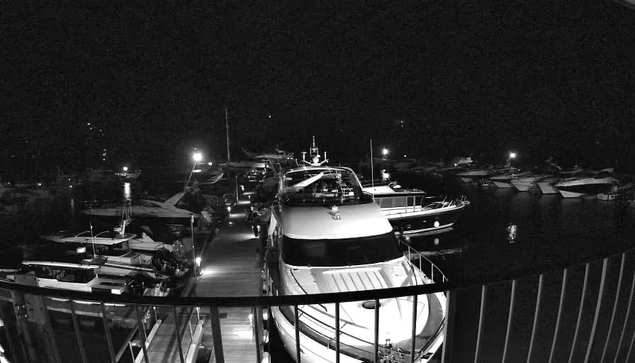 A nighttime scene of a marina with several boats docked. The image is in black and white, featuring a large white boat in the foreground and various smaller boats in the background. Soft lights illuminate parts of the marina, reflecting on the water's surface. The atmosphere appears calm and quiet, with no visible people.