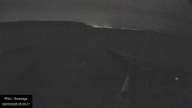 A dimly lit landscape at night showing a dark, blurred coastline with faint lights in the distance. The foreground features indistinct outlines of objects, possibly part of a boat or structure. The overall scene is mostly black with low visibility.