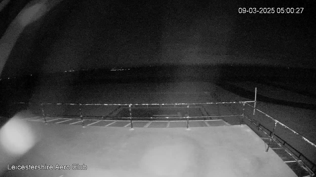 A dark scene captured from a webcam at the Leicestershire Aero Club. The image shows a flat surface, likely a runway or platform, with a railing in the foreground. In the background, faint lights are visible in the distance, suggesting a horizon or distant structures, but it is predominantly dark. The timestamp indicates the image was taken early in the morning.