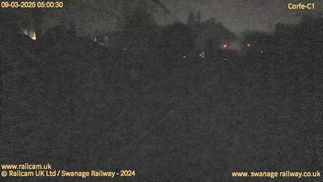 A dimly lit scene captured by a webcam, likely set in an outdoor environment during the early morning hours before dawn. The image is predominantly dark, with indistinct shapes of buildings and trees appearing against the night sky. Soft, warm light glows from a few sources in the background, possibly windows or street lamps. There are faint red lights visible, suggesting the presence of signal indicators or other equipment. The overall atmosphere is quiet and still, typical of early morning before the day begins.