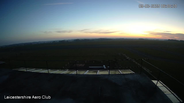 A view from a webcam at Leicestershire Aero Club, showing a wide open space during dawn. The sky is transitioning from dark to light, featuring hues of blue and soft orange as the sun rises in the horizon. Below, an expanse of grassland is visible with faint outlines of fences or pathways, indicating an airfield. The foreground includes a rooftop ledge with a few panels and some structural elements, suggesting the perspective is from a high vantage point.