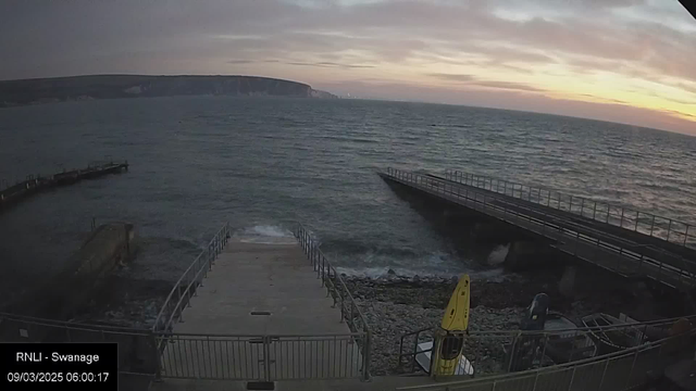 A view of the sea at dawn, with gentle waves lapping against a rocky shoreline. In the foreground, a concrete ramp leads down to the water, flanked by railings. On the right side, a long wooden pier extends over the water, while a yellow kayak rests near the ramp. The sky is filled with soft clouds and a gradient of colors from pale orange to gray, indicating the early morning light. The horizon features a distant cliff, partially shrouded by mist.