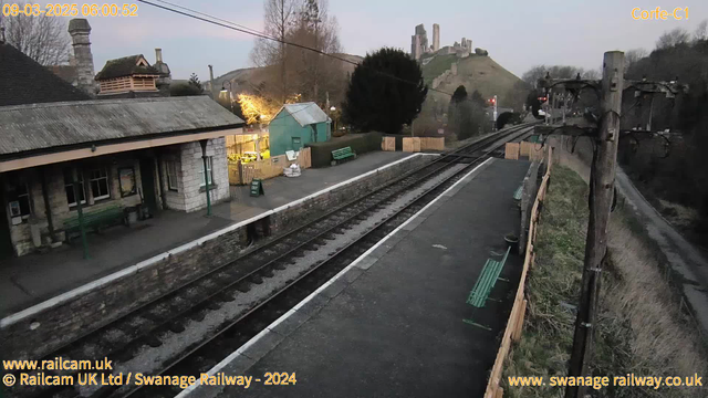 A view of a quiet train station at dawn, with a stone platform and two sets of railway tracks leading away into the distance. On the left, there is a small, stone building with a sloped roof and a green bench in front. Opposite the building, there is a green shed and more benches along the platform. In the background, a hill rises, with the remnants of a castle partially visible at the top. Trees are scattered around the scene, and the sky is lightening with the first light of day.