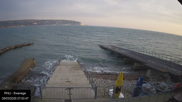 A coastal scene is depicted with a view of the sea and rocky shore. In the foreground, there is a concrete ramp leading down to the water, bordered by a metal railing. To the right, a wooden pier extends over the water, with several boats parked on the shore. A few colorful kayaks in yellow and blue are positioned nearby. The water appears calm, and the sky is cloudy with a soft gradient, indicating early morning light. In the background, cliffs can be seen rising above the sea.