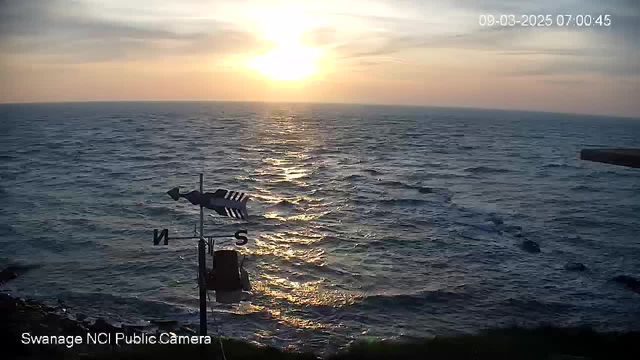 A view of the ocean at sunrise, with golden light reflecting on the water's surface. The horizon features soft clouds and a gradient of colors from yellow to blue. In the foreground, a weather vane with arrows points southward against a backdrop of waves. The scene is tranquil, capturing the beauty of early morning by the sea.